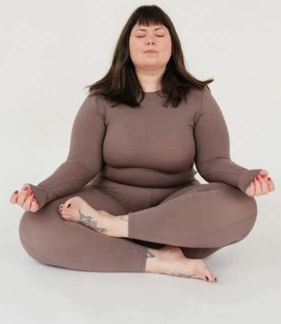 FULL-SIZED GROWTH MINDSET PICTURE OF A YOUNGER GIRL WITH SHOULDER LENGTH BROWN HAIR WITH A BANG IN A GRAY WORKOUT OUTFIT DOING MEDITATION INDIAN STYLE.