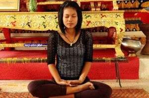 GIRL SITTING IN INDIAN STYLE ON THE FLOOR IN THE SPIRITUAL HEALING PROCESS IN MEDITATION. SHE WEARS A BLACK THREE QUARTER SLEEEVE TOP AND PANTS OUTFIT. BACKGROIND ID YELLOW AND RED COLLORS.