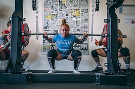 HOW HORMONAL CHANGES AFFECT WEIGHT LOSS = WOMEN AT A GYM LIFTING WEIGHTS. SHW IA WEARING A BLUE T-SHIRT THAT READS BACON