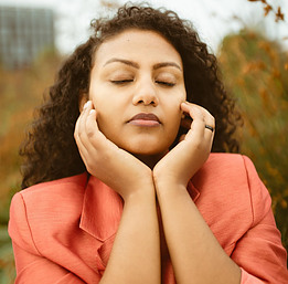 How to Balance Cortisol Levels -Women with Black Curly hair holding her face with her eyes closed wearing a coral jacket