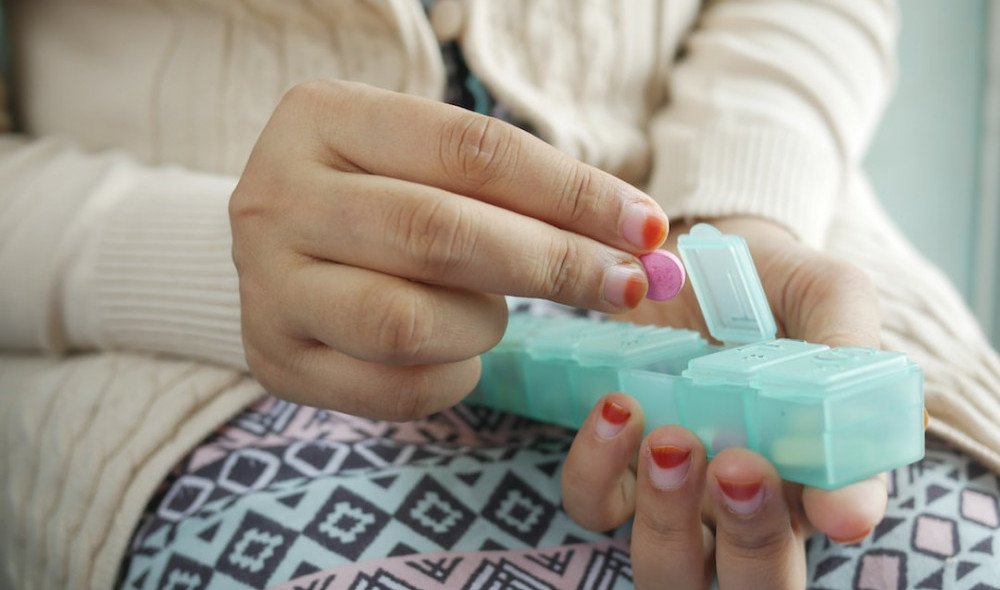 How to Lower Cortisol Levels Someone in a white sweater and printed skirt is taking a pill from her pill pac