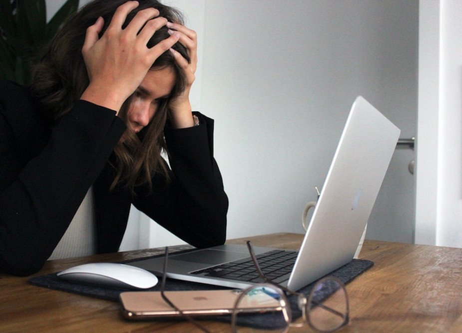 How to Regulate Cortisol Levels - Lady in a black, long sleeve jacket  is sitting at a table in front of her laptop with her hands holding the top of her head.