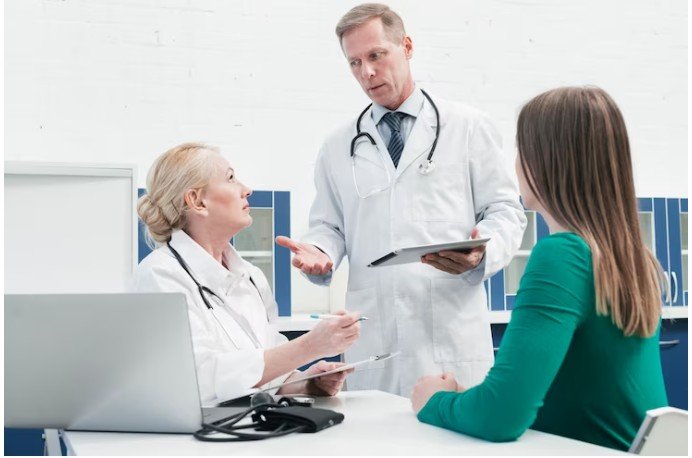 DOCTOR STANDING IN A WHITE LAB COAT WITH STETHOSCOPE AROUND HIS NEXT. HE'S TALKING TO ANOTHER DOCTOR WHO IS SITTING DOWN