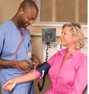 HIGH BLOOD PRESSURE TAKEN BY HER DOCTOR AS A LADY IN  A PINK  TOP SITS IN CHAIR.