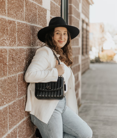 PLUS SIZE MODEL LEANING AGAINST BRICK WALL WEARING A WIDE BRIMMED BLACK HAR, WHICH SWEATER AND LIGHT BLUE JEANS