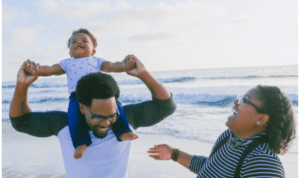 A FATHER IS OMPORTANT TO HELP THEIR CHILD DEVELOP SOCIALLY. A FATHER HAS HIS CHILD ON HIS SHOULDERS AS MOTHER LOOKS ON AT THE BEACH