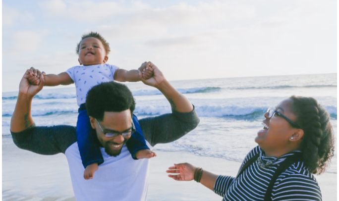 HOW FATHER'S DAY BECAME A NATIONAL HOLIDAY> A FATHER IS IMPORTANT TO HELP THEIR CHILD DEVELOP SOCIALLY. A FATHER HAS HIS CHILD ON HIS SHOULDERS AS MOTHER LOOKS ON AT THE BEACH