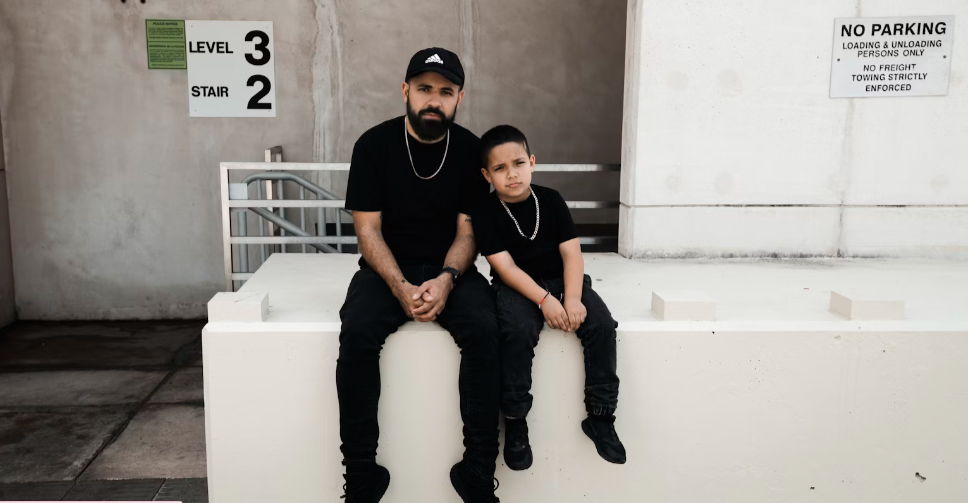 FATHER AND SO BOTH DRESSED IN BLACK WITH A GOLD NECKLACE AT A JOBSITE. TEACHES HIS SON DISCIPLINE