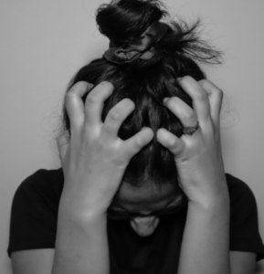 YOUNG WOMAN WITH BLACK HAIR HEAD FACED DOWN HOLDING HER HANDS ON HER HEAD WITH HER FINGERS IN HER HAIR IN FRUSTRATION. WEARING A BLACK SHORT-SLEEVED TOP