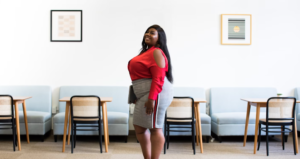 YOUNG ENTREPRENEUR IS PROUD AS SHE STANDS IN HER OFFICE WEARING A RED LONG SLEEVED TO WITH SHOULDERS CUT OUT AN A GRAY SKIRT OVER THE KNEE. SHE COULDBE MORBIDLY OBESE