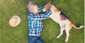 LADY WEARING BLUE JEANS AND A BLUE WHITE AND GREEN PLAID SHIRT HUGGING HER MEDIUM SIZED DOG ON THE GRASS. 