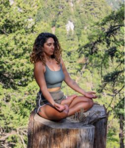 YOUNG LADY OUTDOORS SITTING ON A LARGE TREE STOMP IN A YOGA PISITION DOING MINDFULNESS PRACTICES