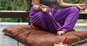 LADY WEARING A BLACK TOP AND PURPLE DRAPED GENIE PANTS WITH HER LEGS AND FEET CROSSED ON A SOFT BROWN MAT DOING MINDFULNESS YOGA