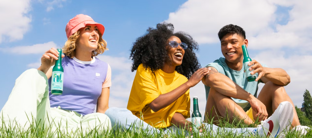 3 FRIENDS FROLICKING OUTDOORS SITTING ON THE GROUND SIPPING NON-A;COHOLIC DRINKS