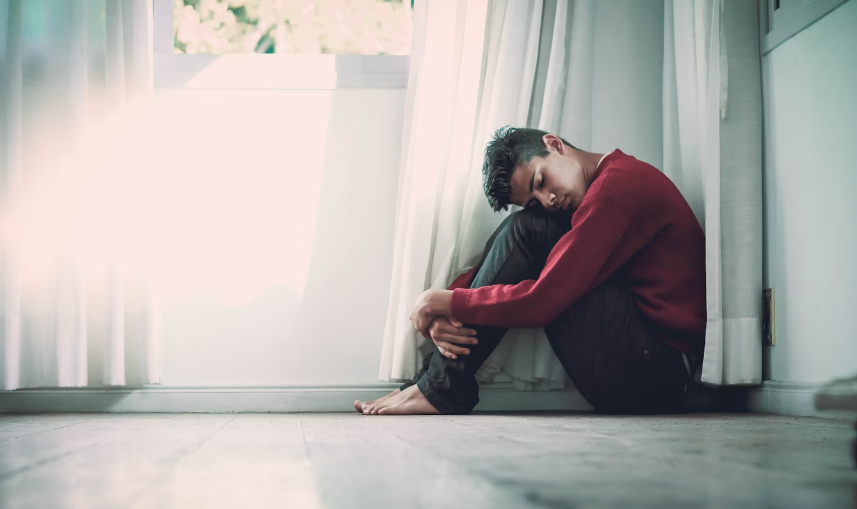 YOUNG MAN SITTING ON FLOOR WITH SEPARATION ANXIETY HEAD DOWN HOLDING HIS KNEES WEARING A LONG-SLEEVED BURGANDY POLO SHIRT