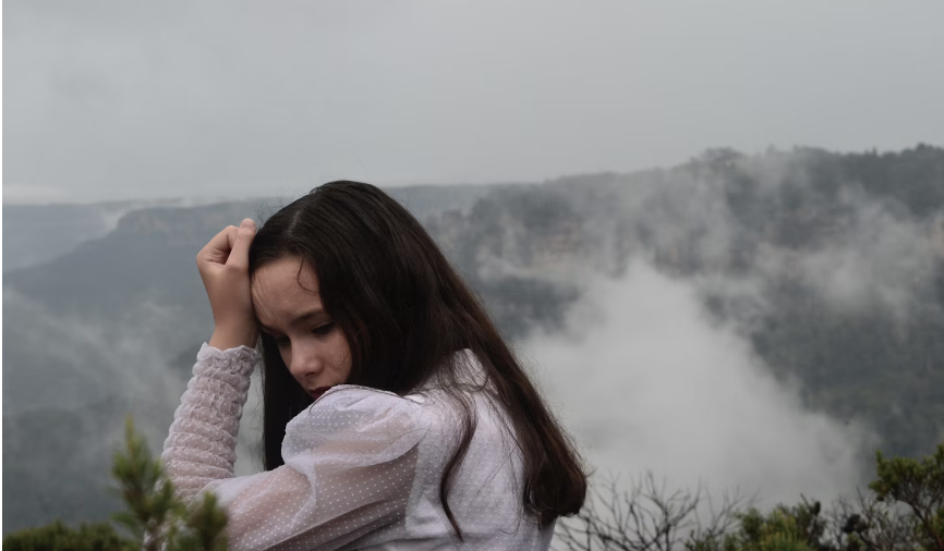 AN ATTRACTIVE YOUNG LADY WITH LONG BLACK HAIR IN LONG SLEEVED WHITE TOP WITH PUFFY SHOULDER LOOKS DOWNWARD HOLDING LEFT HAND TO RIGHT SIDE OF HER HEAD